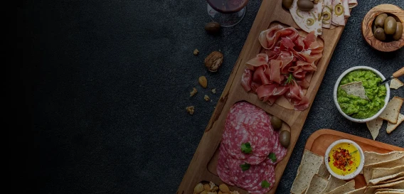 Charcuterie board with cured meats, dips, olives, and crackers on a dark surface.