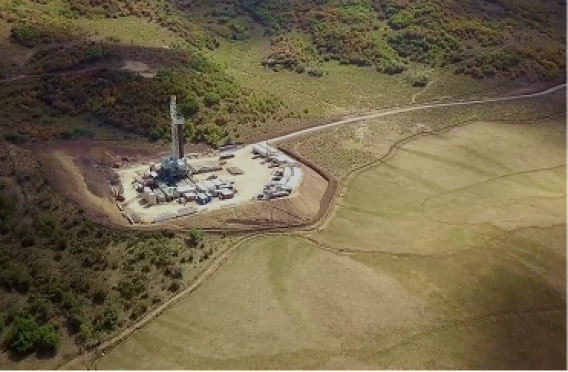 - Industrial site with tall drilling rig, storage units, and dirt roads amid grassy hills.