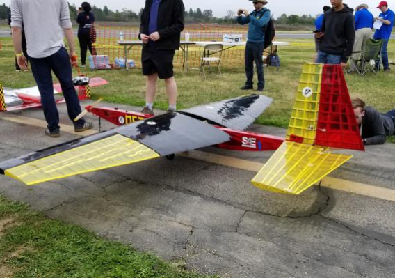 A large remote-controlled aircraft sits on a runway with people standing nearby.