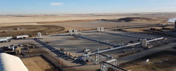 Aerial view of an industrial processing pad with tanks, piping, and vehicles in a rural setting.