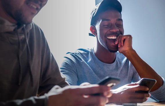 Two young men smiling and looking at smartphones while sitting together indoors.