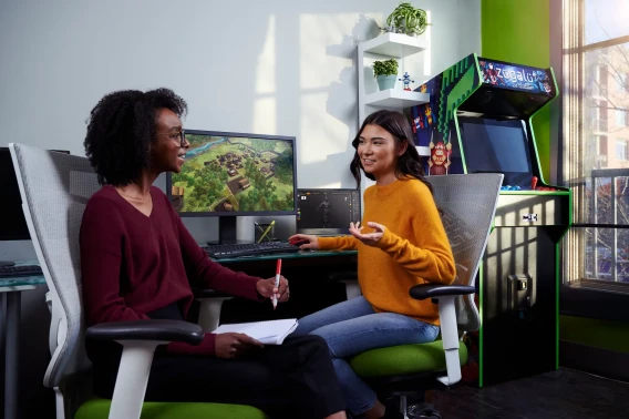 Two people talking in a game studio with a computer monitor and arcade cabinet in the background.