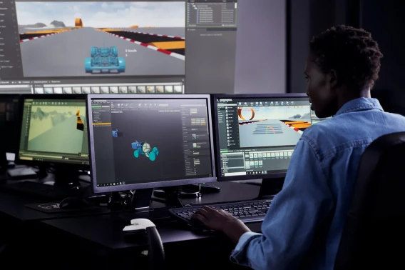 Developer working at multiple monitors with Unreal Engine open, building a racing game environment.