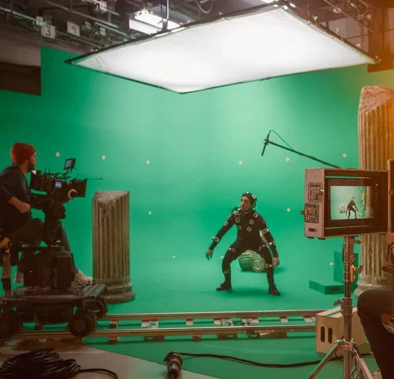 Performer in a motion capture suit stands on a green screen film set with cameras, lights, and monitors.