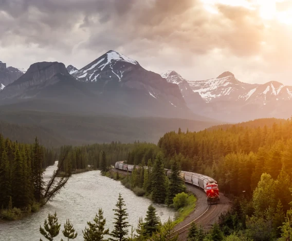 Red train curving beside a river through a forested mountain landscape at sunset.