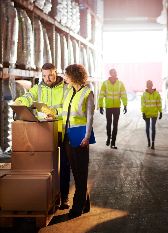 Warehouse staff in safety vests reviewing shipment records on a laptop beside stacked boxes.