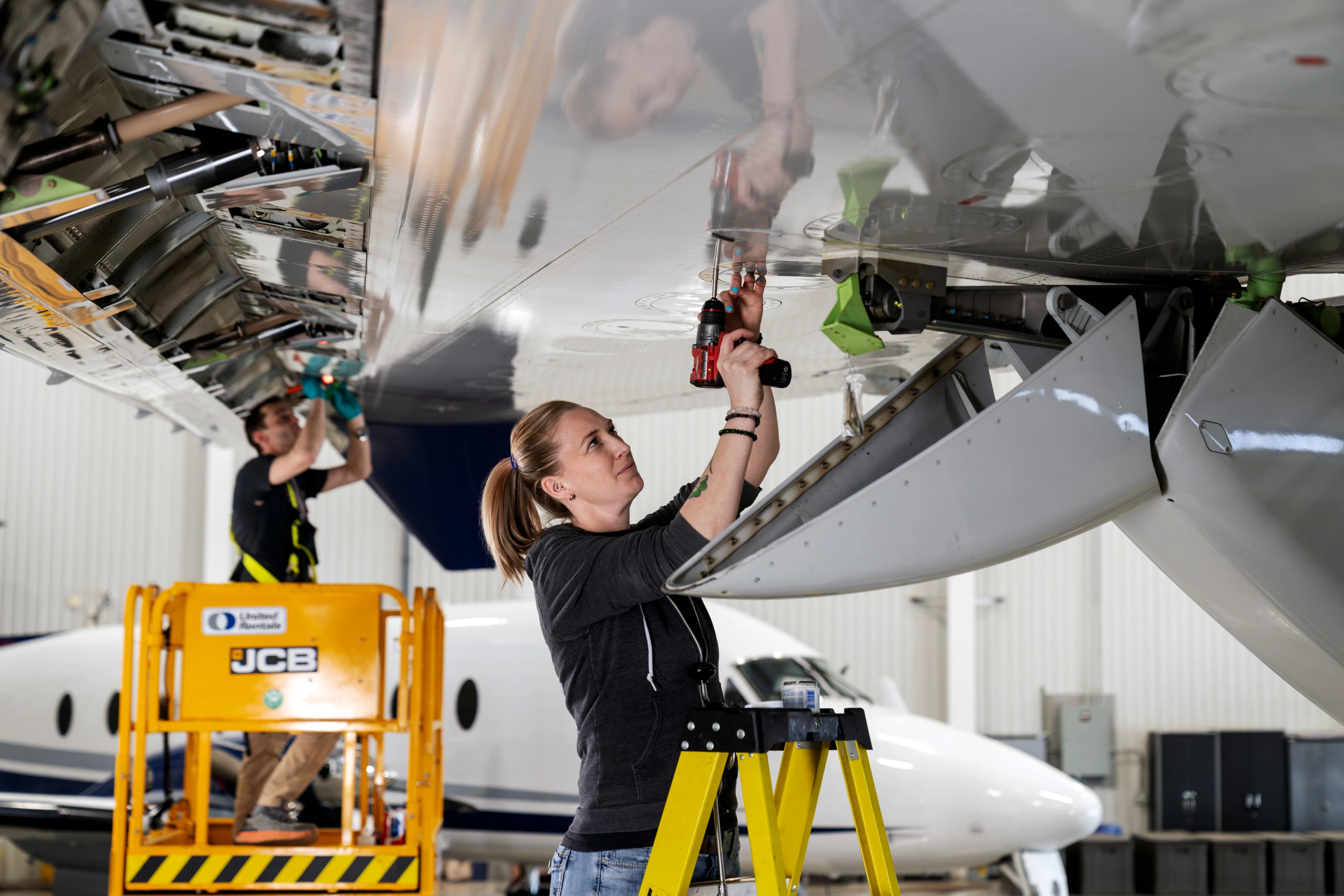 Two technicians perform maintenance underneath an aircraft inside a hangar.