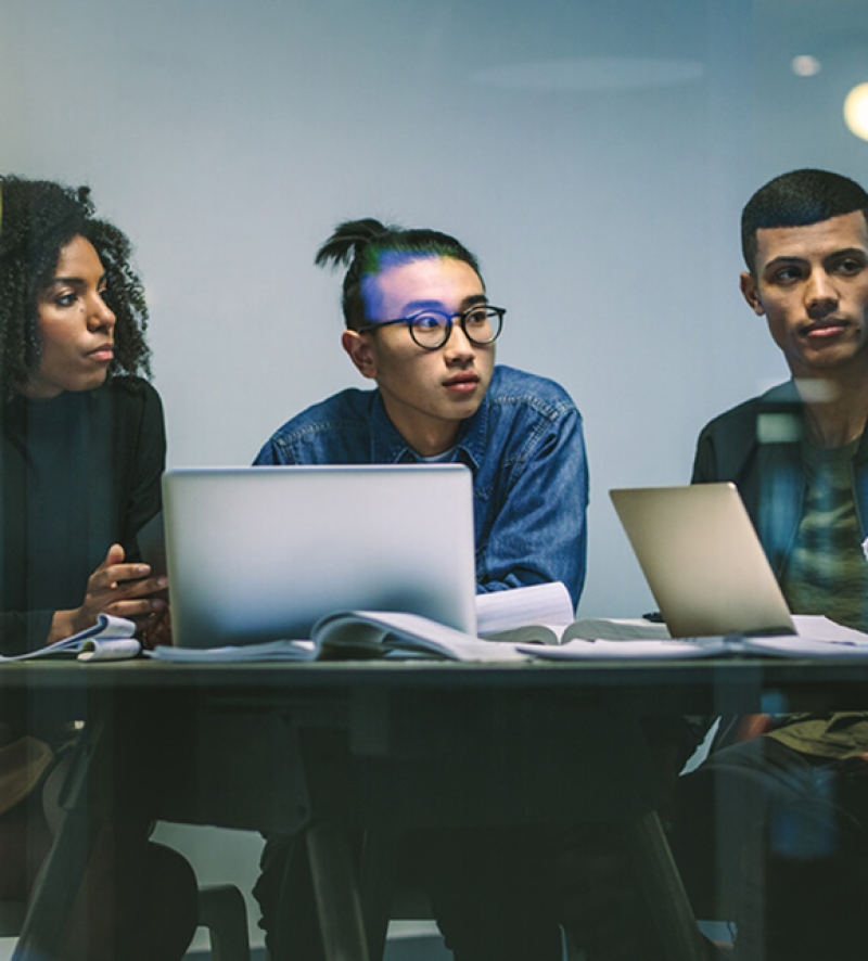 Three professionals seated at table with laptops and papers, engaged in discussion in collaborative workspace.