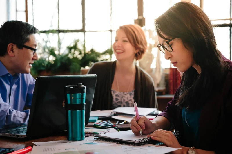 Three students studying together at a table with notebooks, a laptop, and coffee mugs.