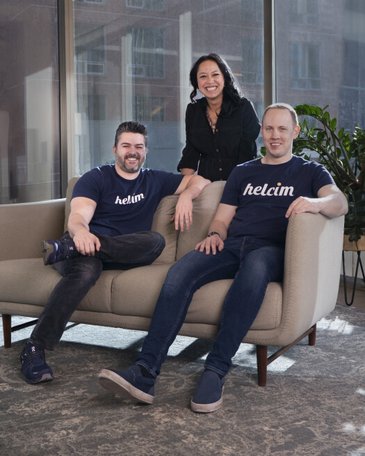 Two men sitting on a couch in black t-shirts reading Helcim with one woman in the middle standing.