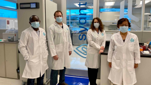 Four people wearing lab coast with masks on standing in front of equipment for a team photo.